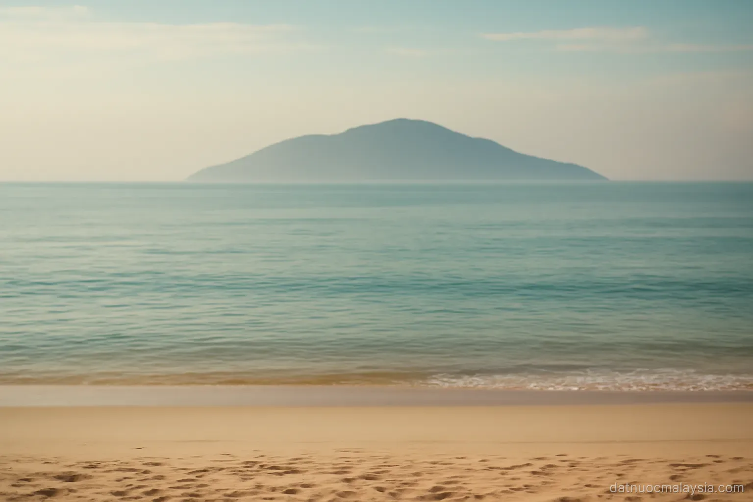 A calm sea with a distant island silhouette under a hazy tropical sky, empty beach in foreground